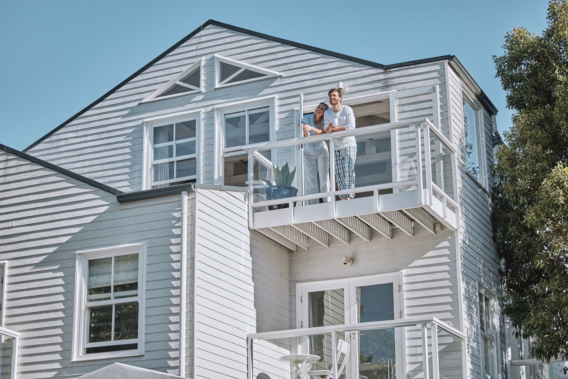 Two people standing on the balcony of a light-colored, modern house on a sunny day.