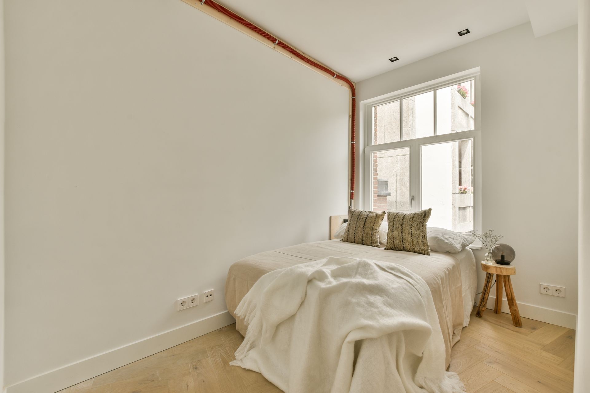 Minimalist bedroom with a beige bed, decorative pillows, wooden side table, and large window.